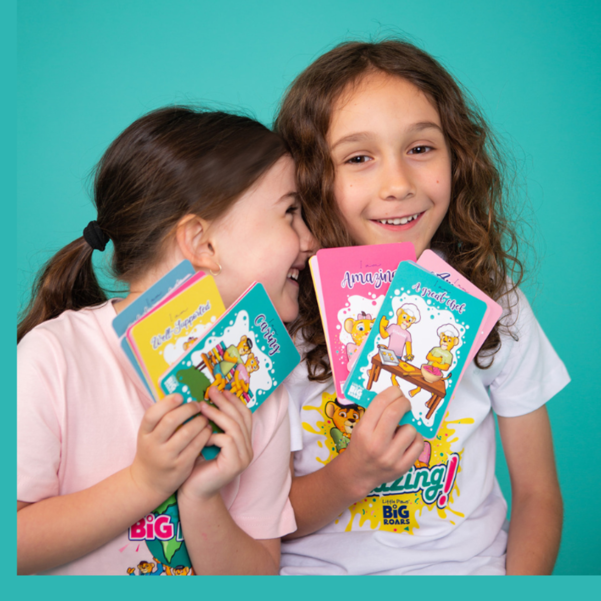 Two children smiling and holding colourful Little Paws Big Roars cards with affirmations such as 'Amazing' and 'A Great Job'. The background is teal, creating a bright and cheerful atmosphere