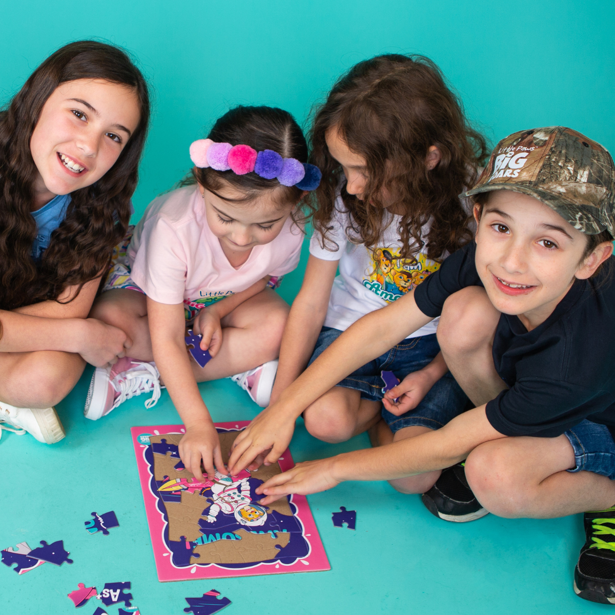 Four children sitting on a teal background, collaborating on assembling a puzzle featuring a vibrant, illustrated image. The children are smiling and engaged, promoting teamwork and creativity