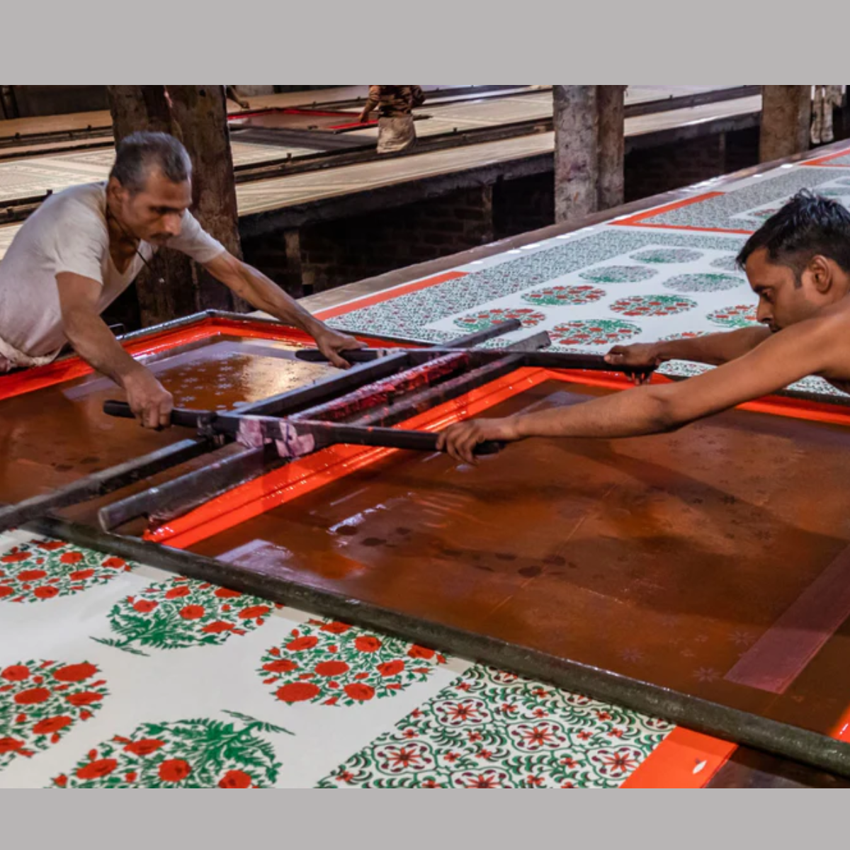 Block Printing Process: Two artisans working collaboratively on a traditional block printing process, using a large frame to print intricate floral and geometric patterns in vibrant colours on fabric. The setting appears to be a workshop with rows of printed textiles.