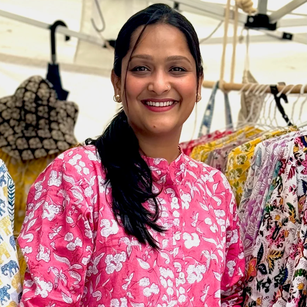 hiHandmade Artisan: A smiling woman wearing a bright pink floral top, standing amidst racks of colourful hand-printed garments. The photo captures a vibrant and cheerful market or boutique setting.