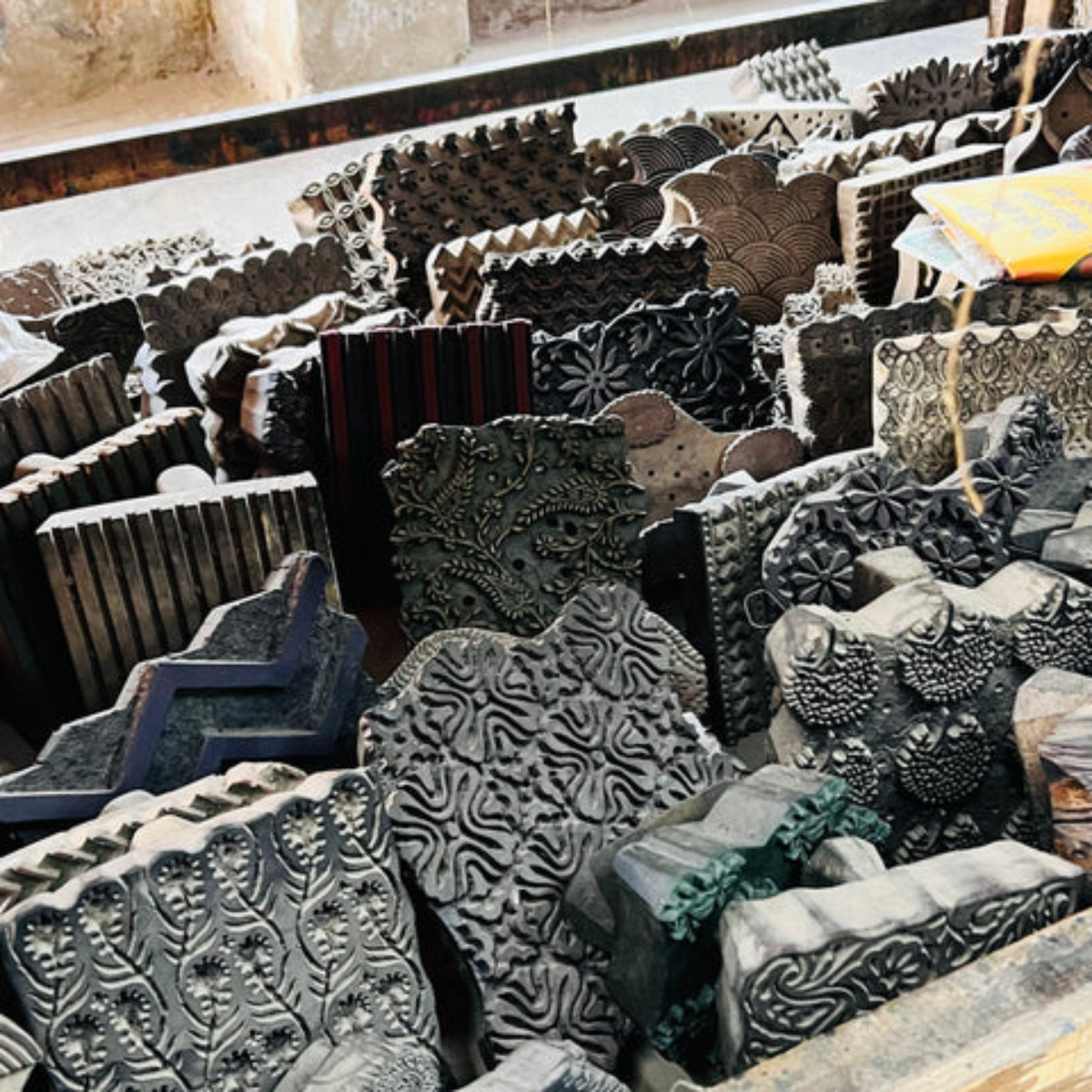 Block Printing Stamps: A collection of intricately carved wooden block stamps, used for traditional fabric printing. The blocks feature a variety of patterns, from floral to geometric, arranged in a workshop-like environment.