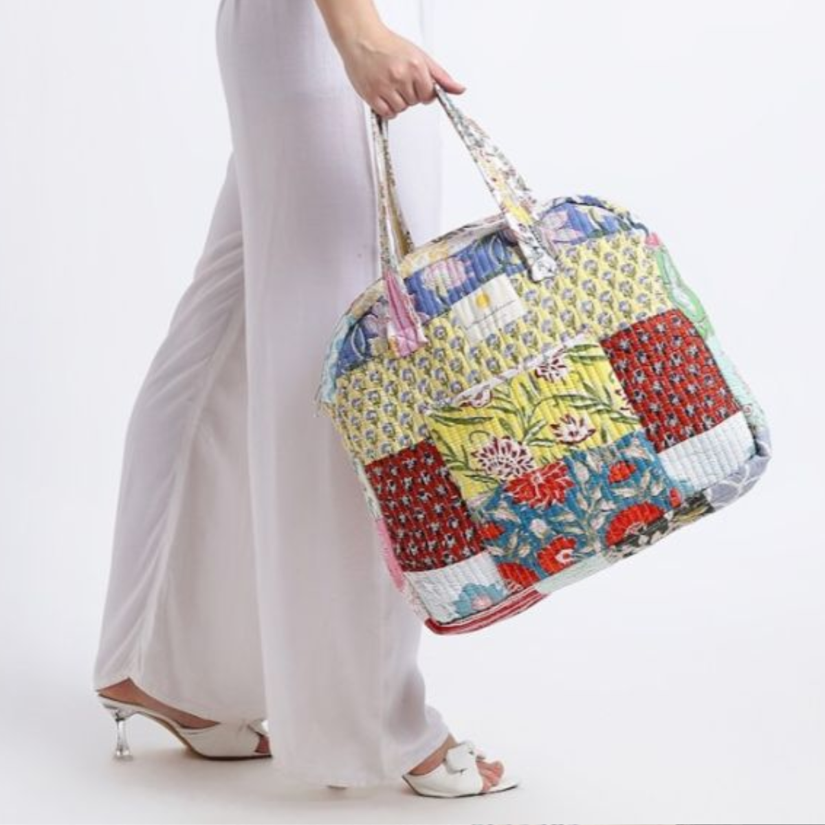 Quilted Bag: A patchwork quilted bag with vibrant floral and geometric patterns in shades of yellow, red, and blue, held by a person wearing white pants and heeled sandals. The bag reflects artisanal craftsmanship and a bohemian style.